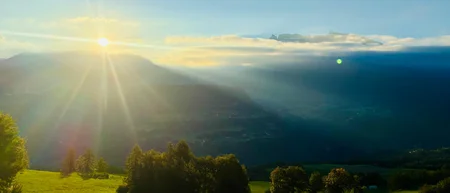 Sonnenaufgang über nebligen Bergen und einem grünen Feld mit Bäumen und Zaun