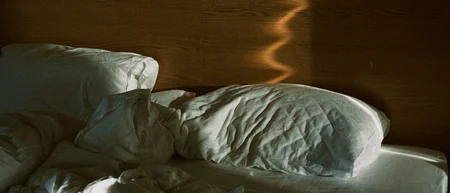 Sonnenlicht zeichnet eine geschwungene Spur auf das Holzkopfteil über einem ungemachten Bett
