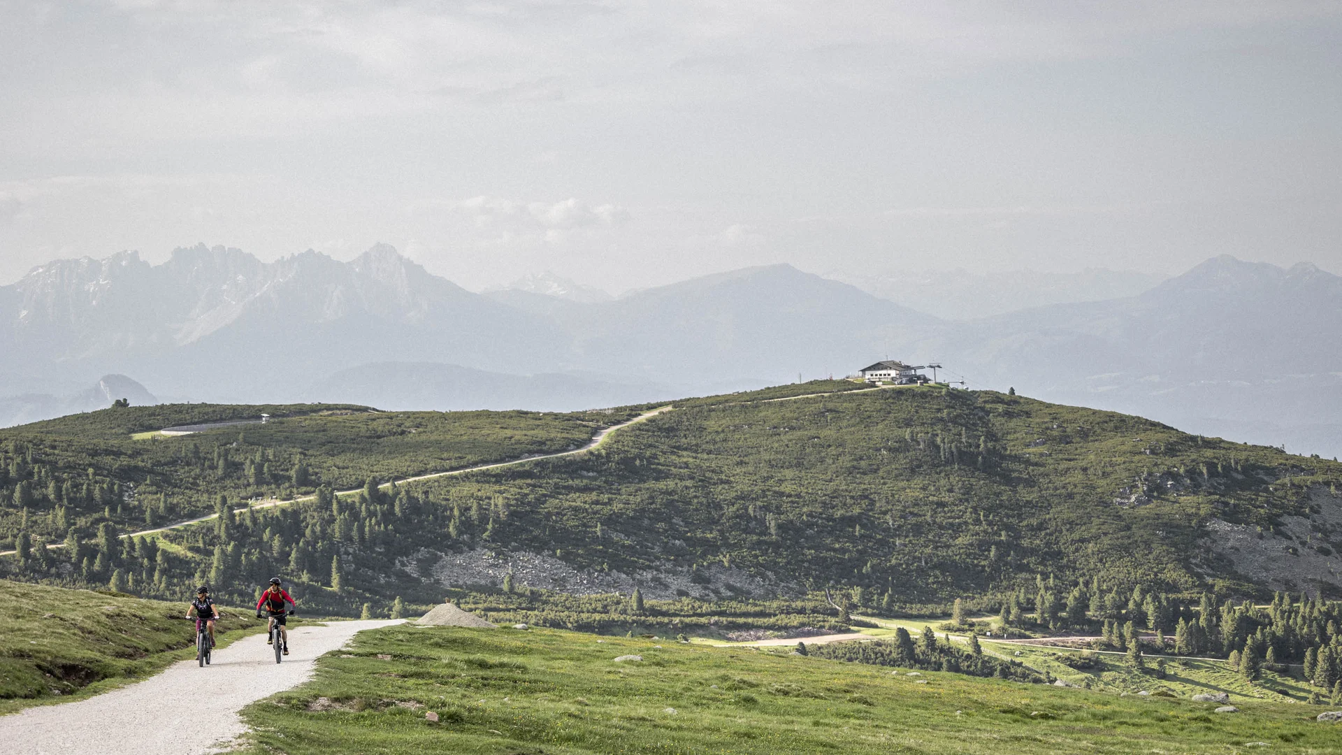 Bilder vom Armonia: Hof am Ritten in Südtirol Zwei Radfahrer auf einem Weg über eine grüne Alm mit Berghütte und Gipfeln im Hintergrund