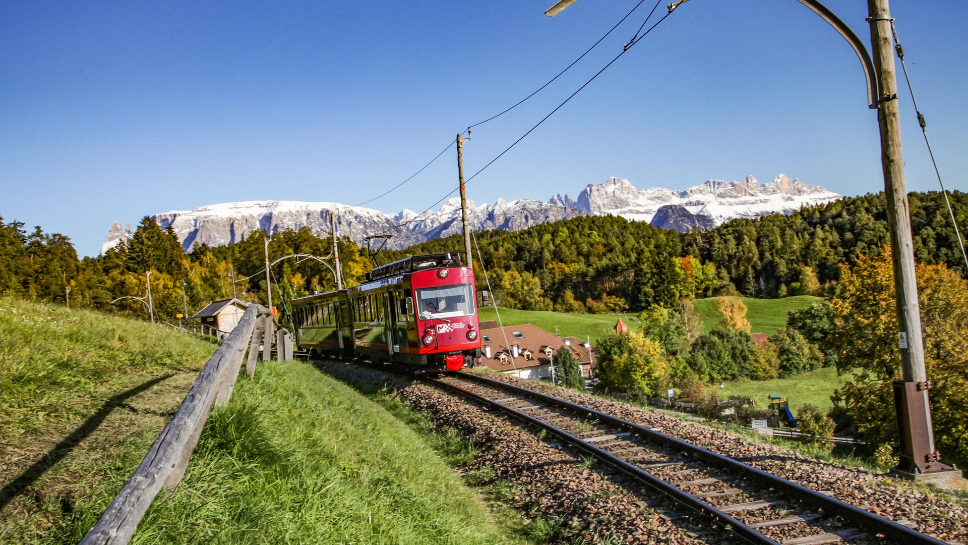 Bilder vom Armonia: Hof am Ritten in Südtirol Rote Bahn auf Gleisen vor grünen Wiesen und schneebedeckten Bergen
