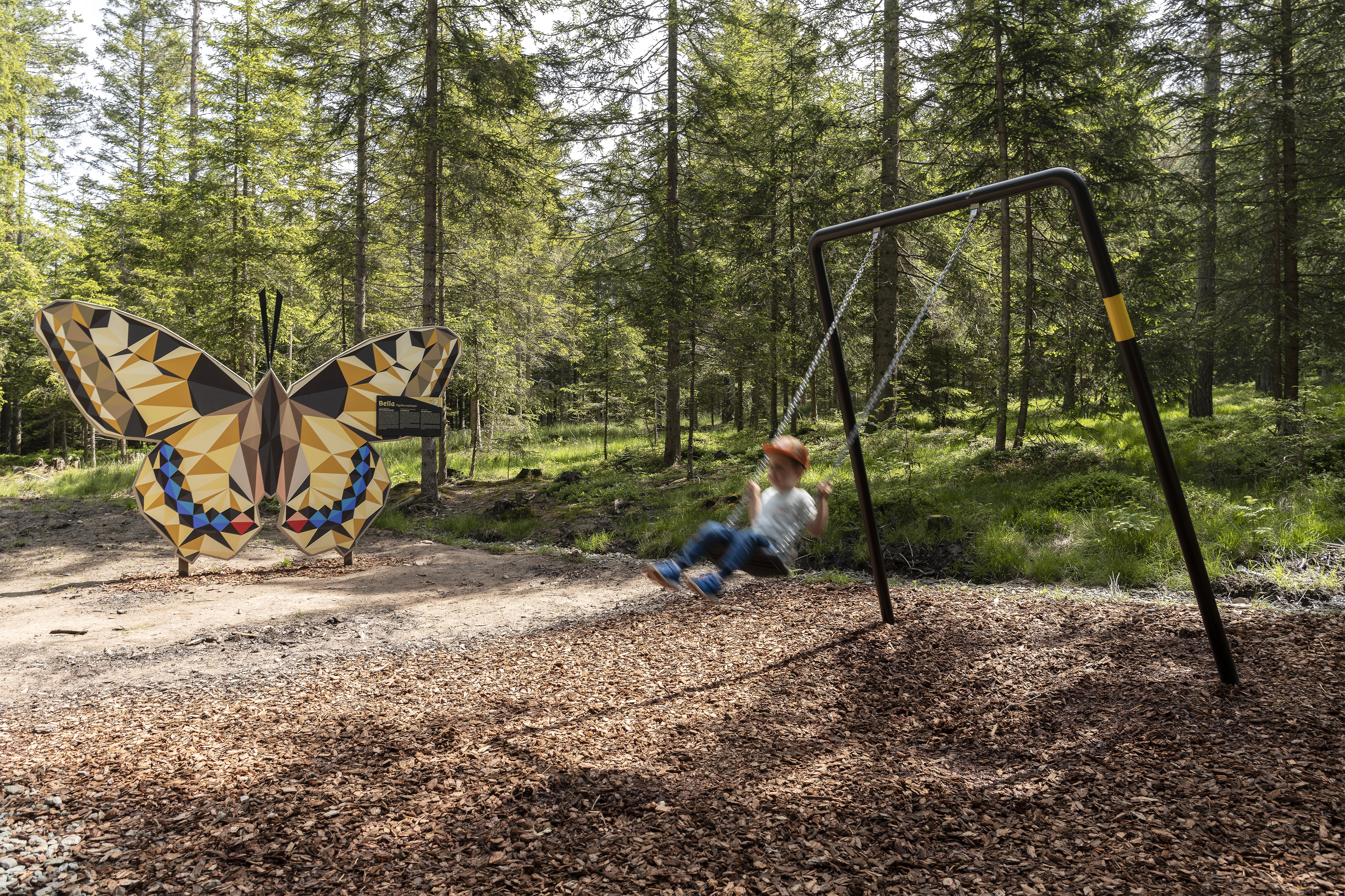 Foto dell’Armonia: agriturismo sul Renon in Alto Adige Un bambino si dondola nel bosco accanto a una grande scultura colorata a forma di farfalla